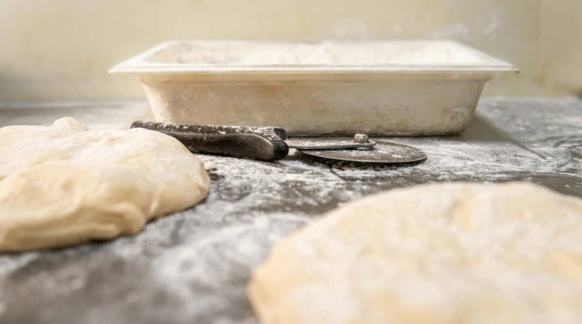 Two mounds of dough on a flour-dusted surface with a pizza cutter and a flour-covered container.