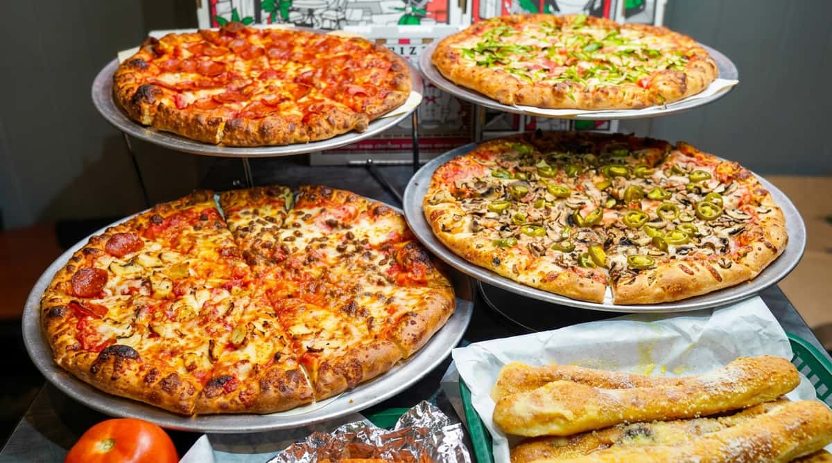 Four pizzas with various toppings (pepperoni, green peppers, sausage, mushrooms/jalapeños), chicken wings, and garlic knots.