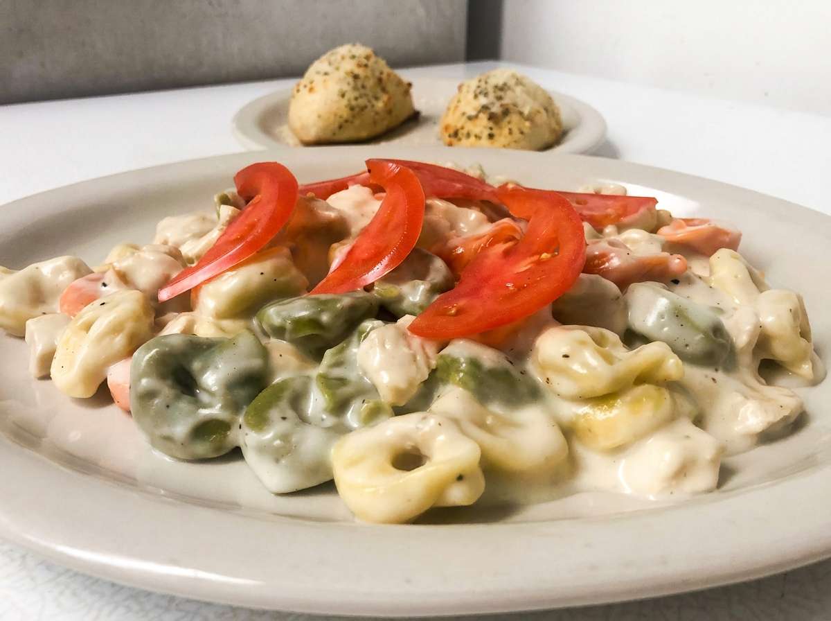 A creamy tortellini pasta dish is served on a white plate, coated in a rich white sauce that generously blankets each piece. The pasta is mixed with tender broccoli florets and topped with thin slices of red bell pepper for a pop of color. In the background, two small garlic knots rest on the plate, lightly browned and sprinkled with herbs, completing this comforting Italian entrée.