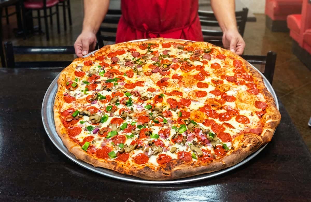 A massive round pizza sits on a large black serving tray, topped generously with melted mozzarella cheese and evenly spaced slices of pepperoni. Fresh chopped herbs—likely parsley or cilantro—are sprinkled across the surface, adding a pop of green color against the rich red sauce and golden cheese. The crust is wide, golden-brown, and slightly crisp around the edges. Behind the pizza, a person wearing a red shirt holds the tray, emphasizing the impressive size and fresh-from-the-oven appeal.