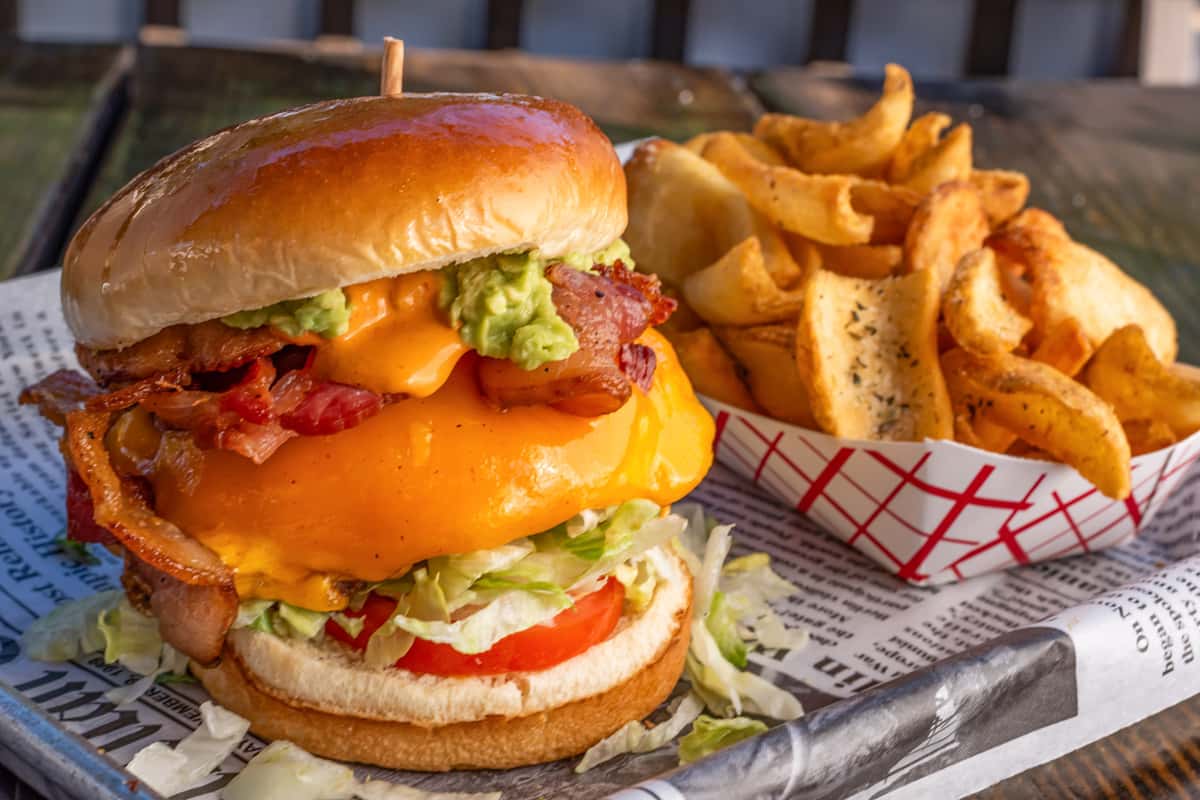 Cheeseburger stacked with bacon, cheddar, guacamole, lettuce, and tomato, served with seasoned curly fries on a metal tray.