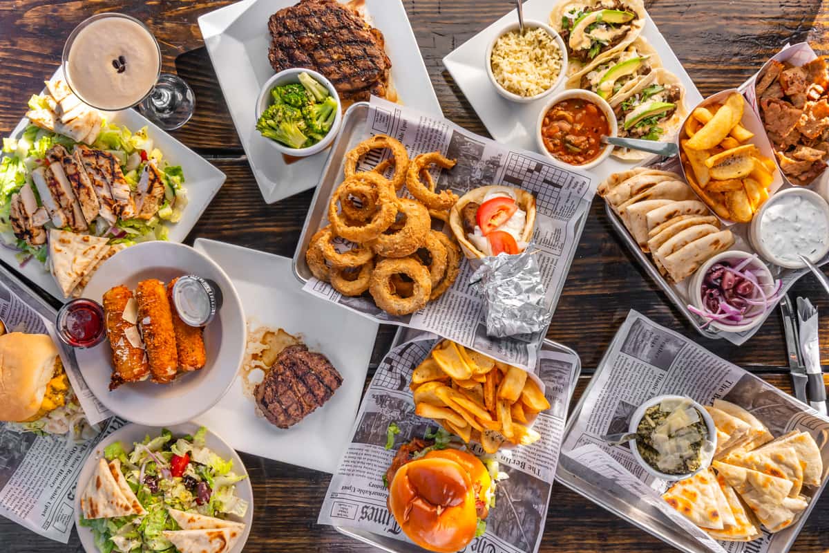 An overhead spread of grilled meats, burgers, tacos, salads, onion rings, pita with dips, and drinks on a rustic wooden table.