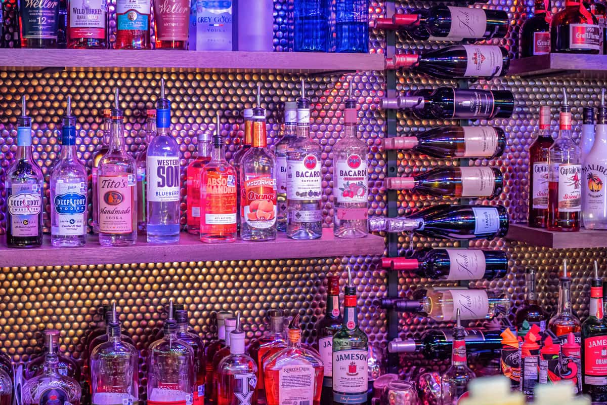 Colorful bar display with shelves stocked with vodka, rum, whiskey, liqueurs, and red wine bottles under moody purple lighting.