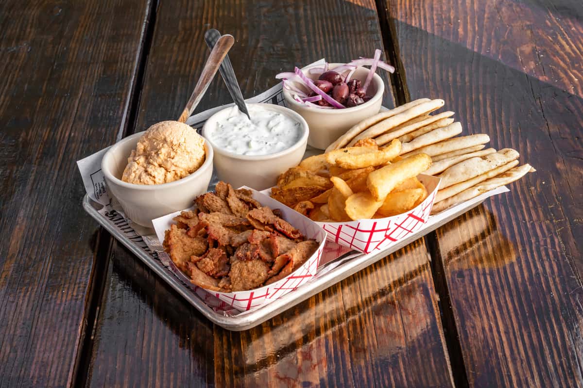 Greek platter with gyro meat, pita bread, seasoned fries, whipped feta, tzatziki, and kalamata olives with red onions on a metal tray.