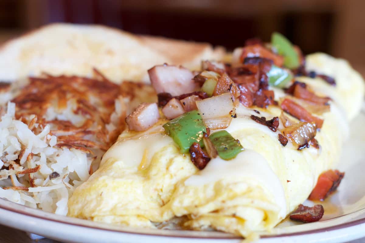 Hearty breakfast plate with a cheesy omelet topped with ham, green peppers, and onions, served with crispy hash browns and toast.