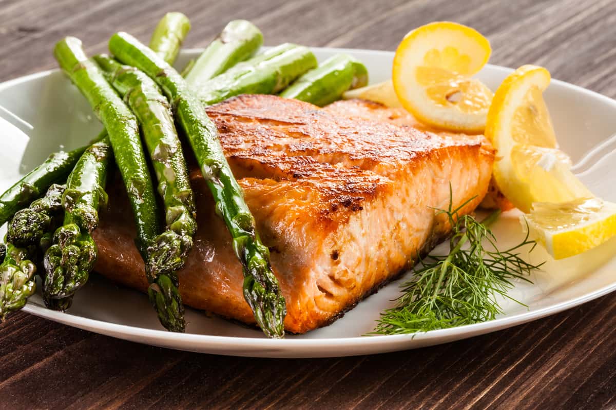 Seared salmon fillet served with grilled asparagus, fresh dill, and lemon slices on a white plate.