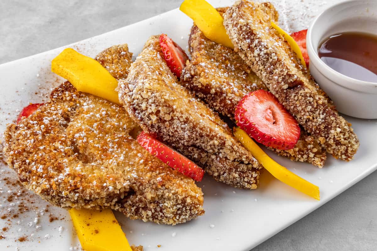 Crunchy-coated French toast dusted with powdered sugar and cinnamon, garnished with fresh strawberry slices and sweet mango strips, served with a side of maple syrup for a vibrant and indulgent brunch plate.