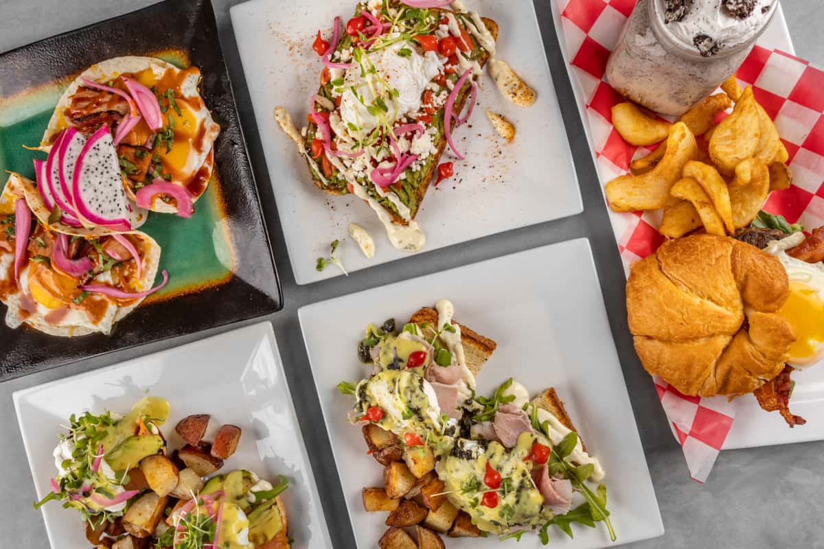 Vibrant brunch spread featuring huevos rancheros with fried eggs and dragon fruit, avocado toast with poached egg and pickled onions, a croissant breakfast sandwich with a sunny-side-up egg and curly fries, two variations of eggs Benedict with home-fried potatoes, and an Oreo milkshake on a checkered tray for a colorful and indulgent brunch experience.