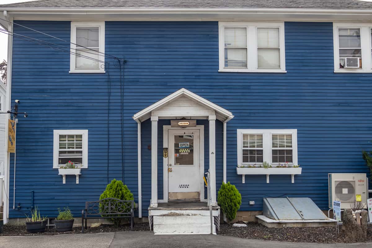 Front entrance of Early Bird brunch restaurant, featuring a cozy white-trimmed doorway with a "We're Open" sign, set against a bold blue exterior with flower boxes, potted plants, and a black bench for a welcoming, small-town café vibe.