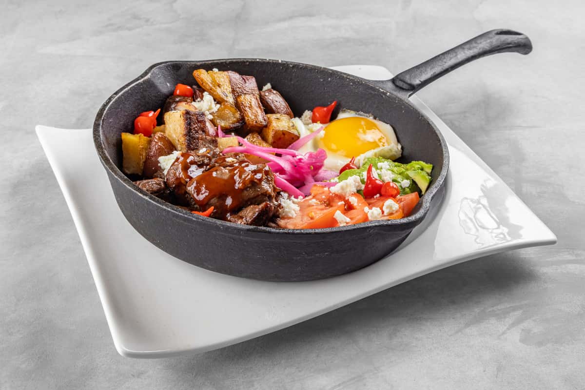 Hearty breakfast skillet served in a cast iron pan, filled with roasted potatoes, braised beef in savory sauce, a sunny-side-up egg, guacamole, diced tomatoes, crumbled queso fresco, and pickled onions for a bold, protein-packed brunch dish.