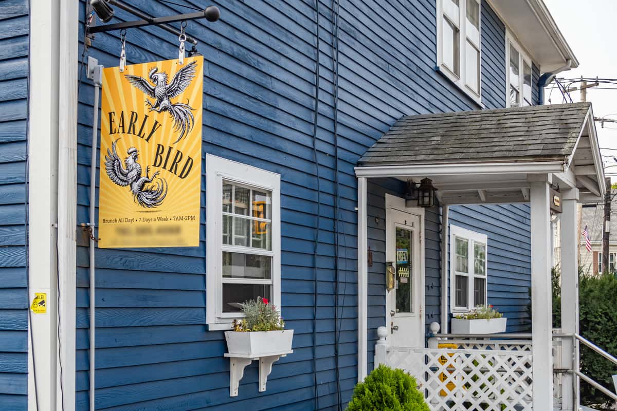 Exterior of Early Bird brunch restaurant featuring a vibrant blue building with white trim, a hanging yellow sign with rooster illustrations, and text reading "Brunch All Day! 7 Days a Week, 7AM–2PM"; white flower boxes with blooming plants enhance the cozy, welcoming atmosphere.