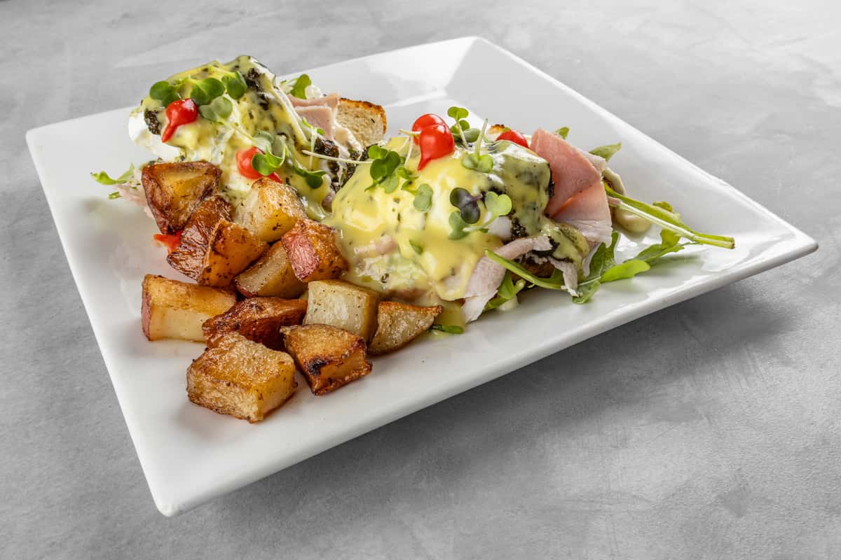Close-up of a gourmet eggs Benedict topped with creamy hollandaise sauce, fresh microgreens, and cherry tomato garnish, served with a side of golden, crispy home-fried potatoes on a white square plate.