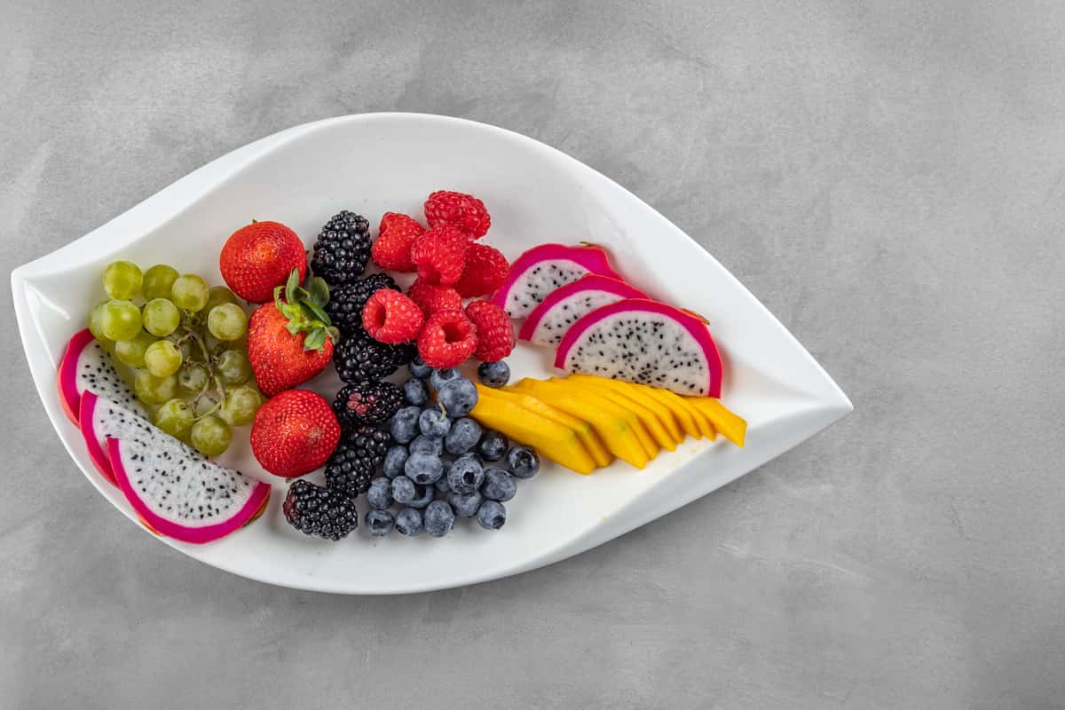 Tropical fruit platter served on a white leaf-shaped dish, featuring vibrant slices of dragon fruit, ripe mango, strawberries, green grapes, blackberries, blueberries, and raspberries for a colorful and refreshing brunch option.