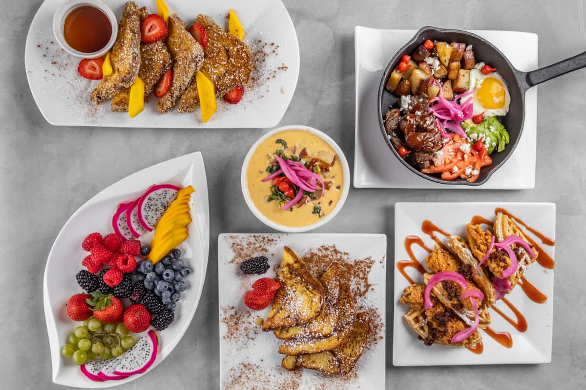 Colorful brunch spread featuring cinnamon-dusted French toast with mango and strawberries, a tropical fruit platter with dragon fruit, berries, and grapes, a creamy queso bowl with pickled onions and herbs, a skillet hash with sunny-side-up egg, guacamole, and roasted potatoes, and crispy chicken and waffles drizzled with spicy sauce and topped with pickled onions.