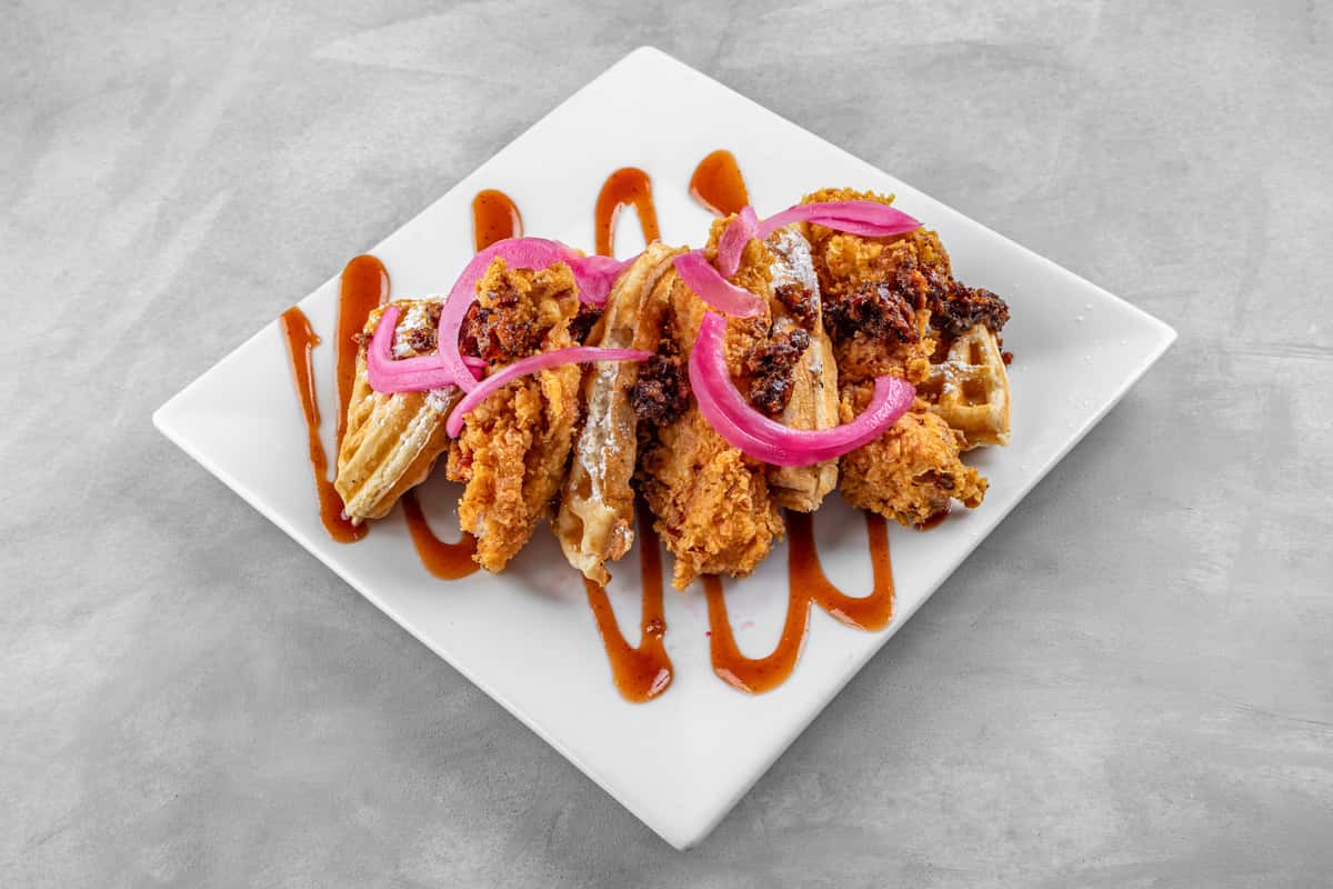 Southern-style chicken and waffles topped with crispy fried chicken, drizzled with a sweet and spicy sauce, garnished with pickled red onions, and served on a white square plate for a bold and flavorful brunch presentation.