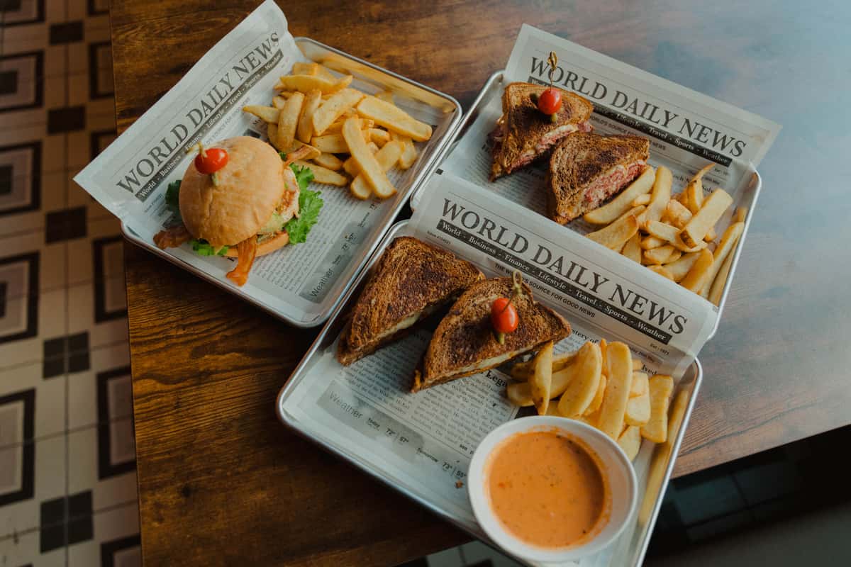 "Assorted Irish pub sandwiches and a burger served with thick-cut fries on newspaper-lined trays, featuring classic pub comfort food on a wooden table.