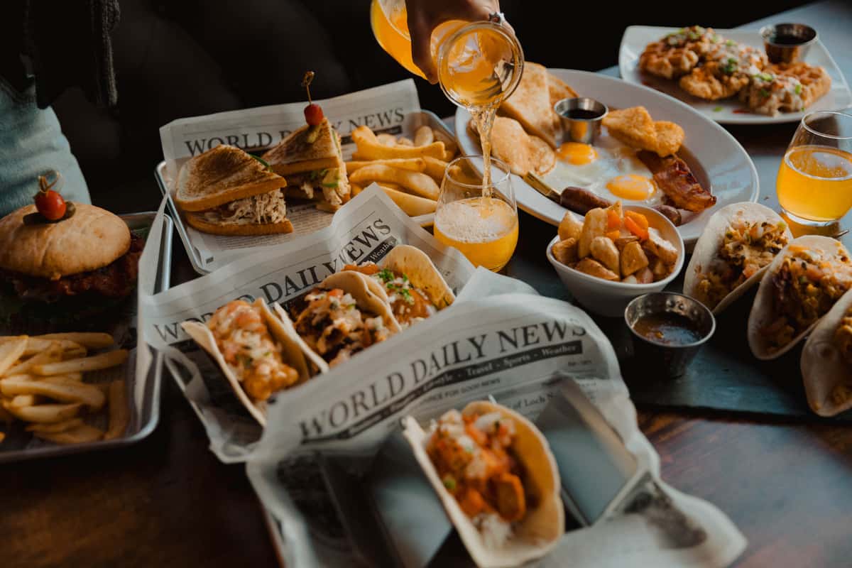 "Irish pub brunch table featuring street-style tacos, toasted sandwiches with fries, burgers, and a full breakfast plate, with fresh orange juice being poured into mimosas.