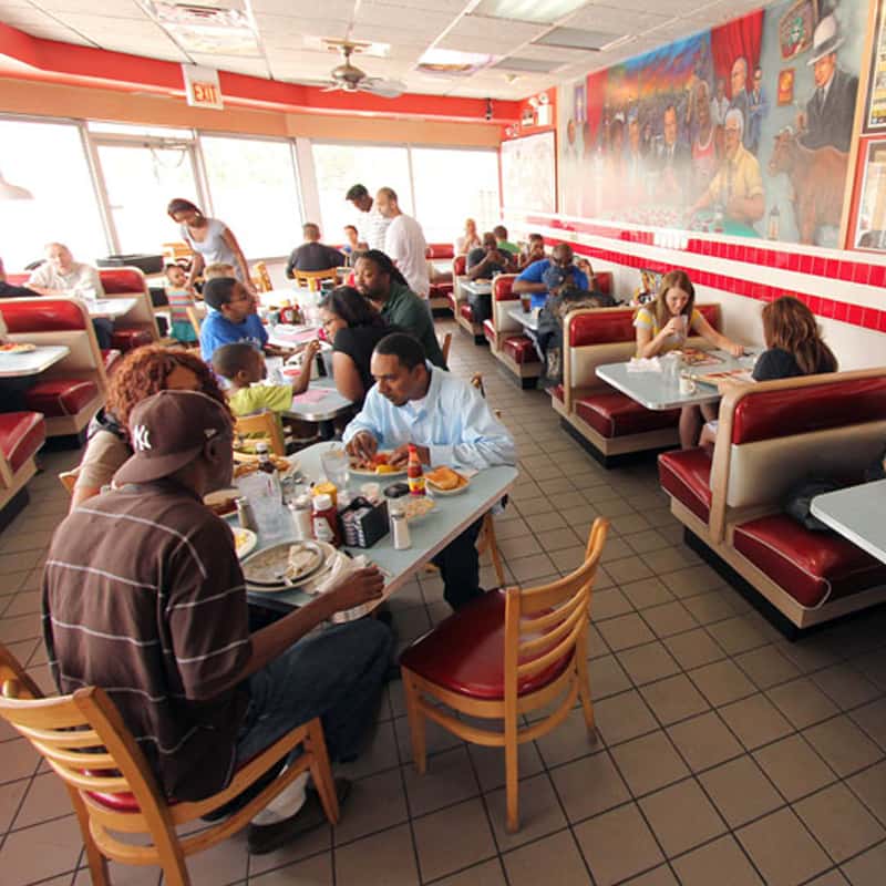 Busy diner scene with red booths and several groups of people eating and talking at tables near large windows and a wall mural.