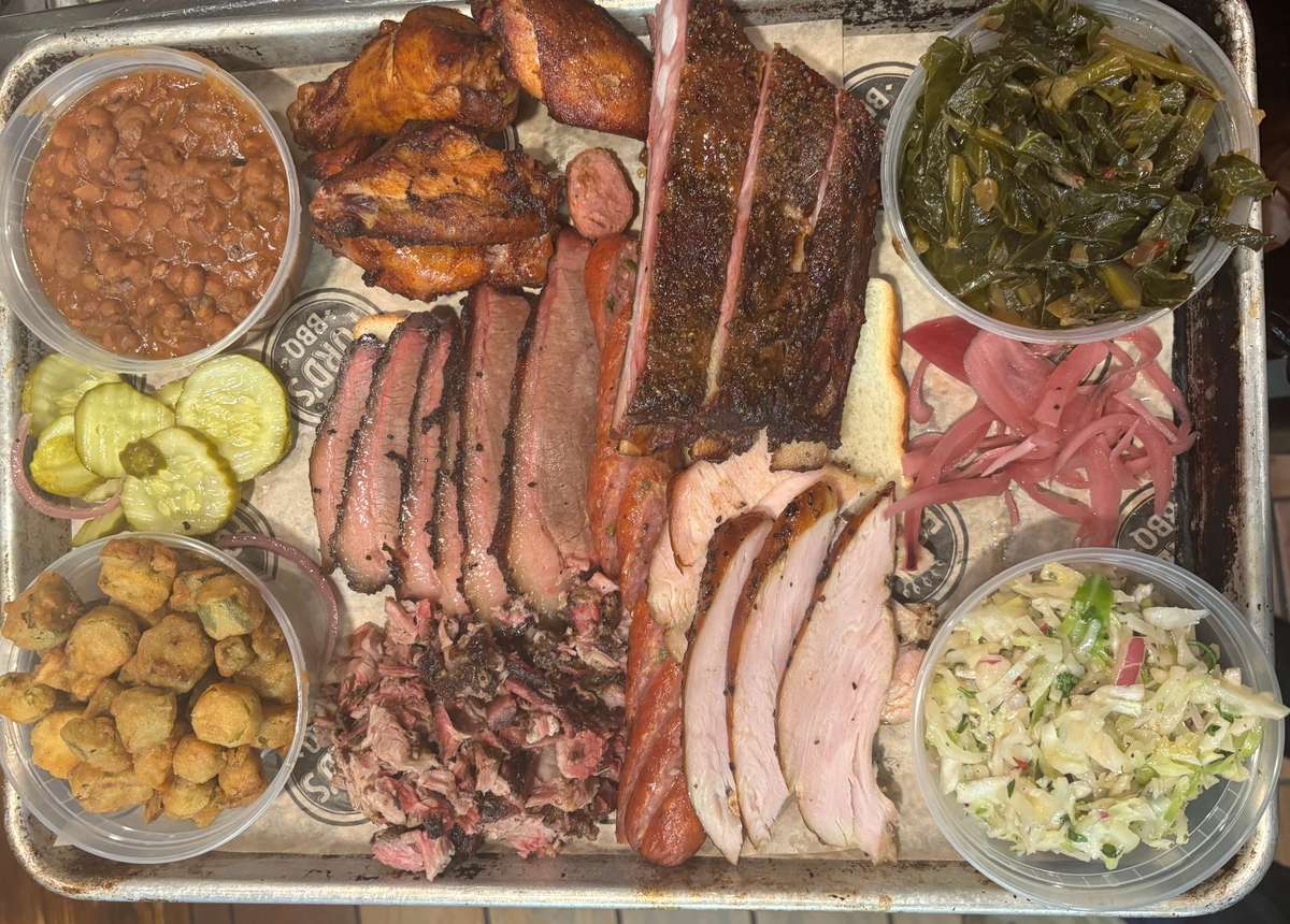 A tray of BBQ meats including sliced brisket, sausage, pulled pork, ribs, and turkey, served with pickles, pickled onions, collard greens, baked beans, okra, cole slaw, and bread.