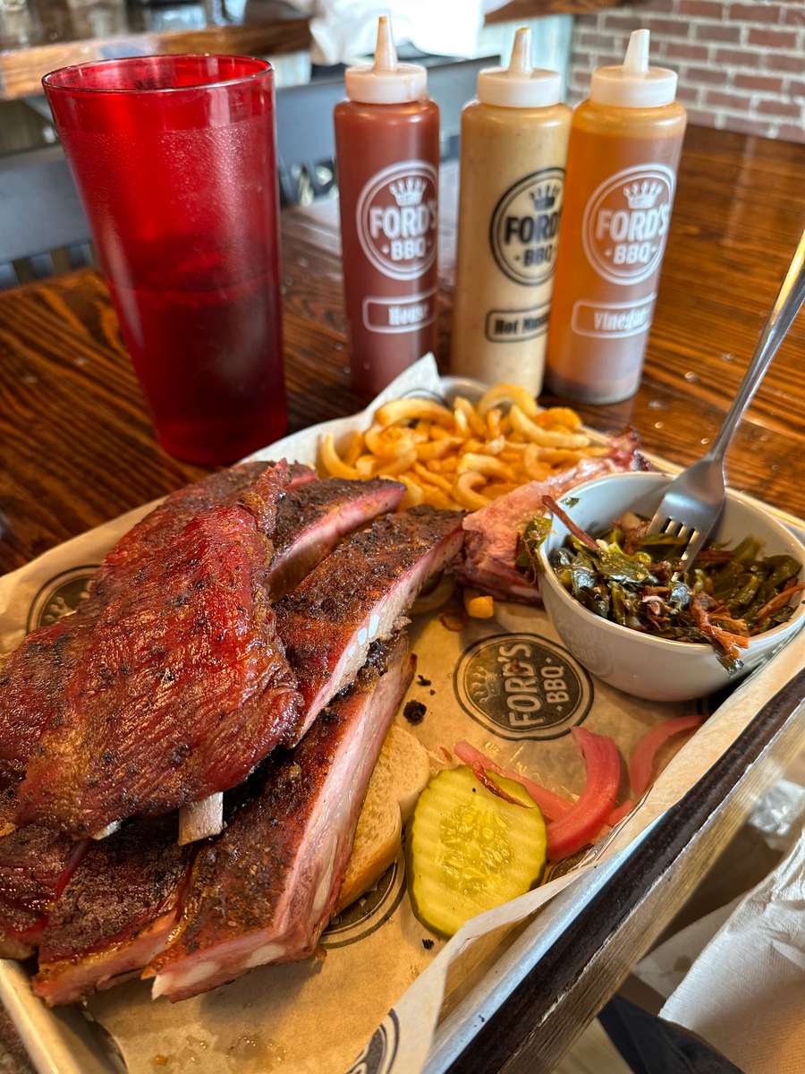 This image features a plate of delicious BBQ ribs served with a side of curly fries and collard greens. The ribs are well-seasoned with a charred crust and accompanied by a slice of bread and pickles. Three bottles of BBQ sauce—likely for customizing flavor—are visible in the background, along with a large glass of drink on the side. The meal is placed on a paper-lined tray with the "Ford's BBQ" branding.