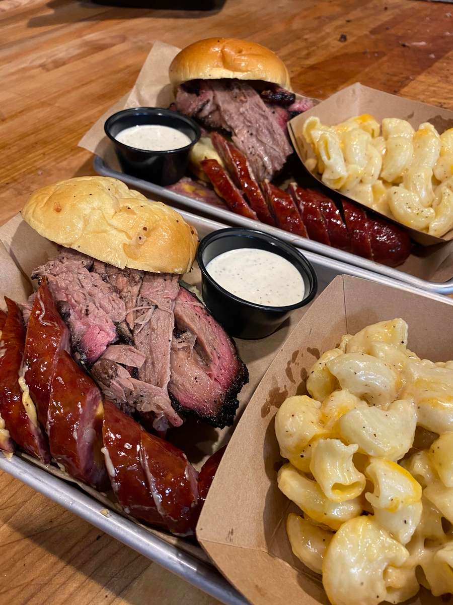 The image shows two BBQ sandwiches, one filled with tender brisket and the other with sliced sausage, paired with sides of creamy macaroni and cheese and dipping sauces.
