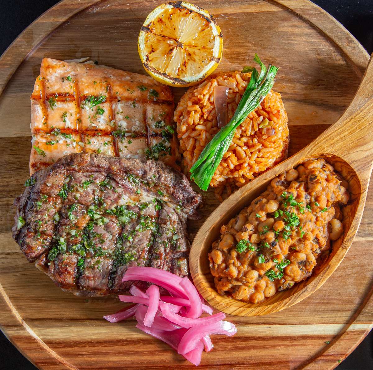 Grilled salmon and steak served with jollof rice, sautéed black-eyed peas, pickled onions, and a grilled lemon on a wooden plate.