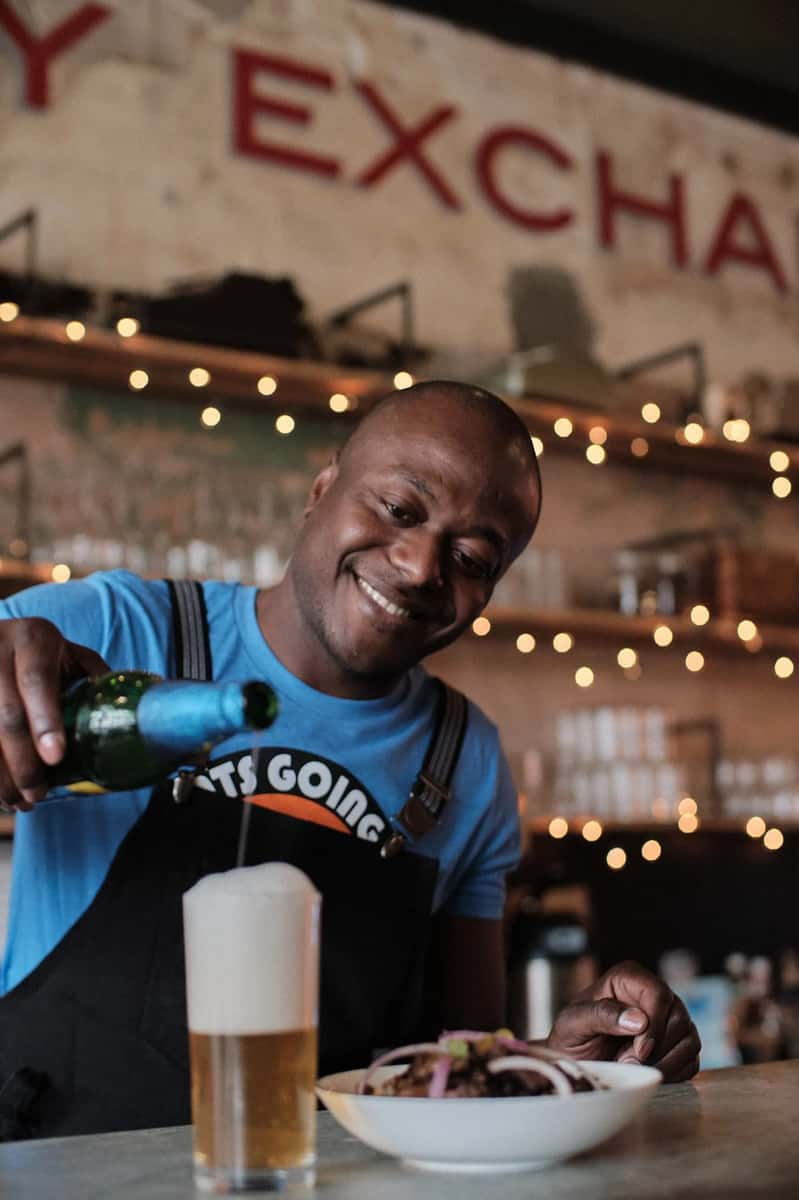 Smiling man in a blue shirt and apron pours a beer beside a bowl of food under string lights in a rustic restaurant setting.