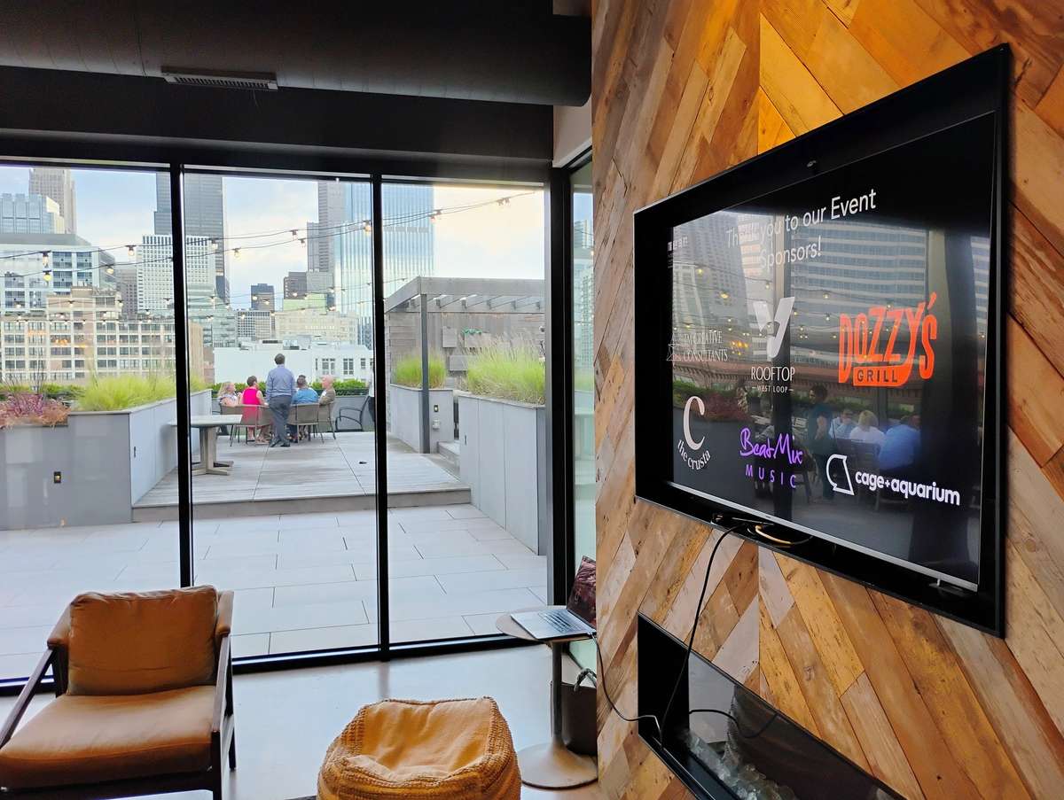 Rooftop lounge with city skyline views, a TV displaying event sponsors, and guests seated at an outdoor table.