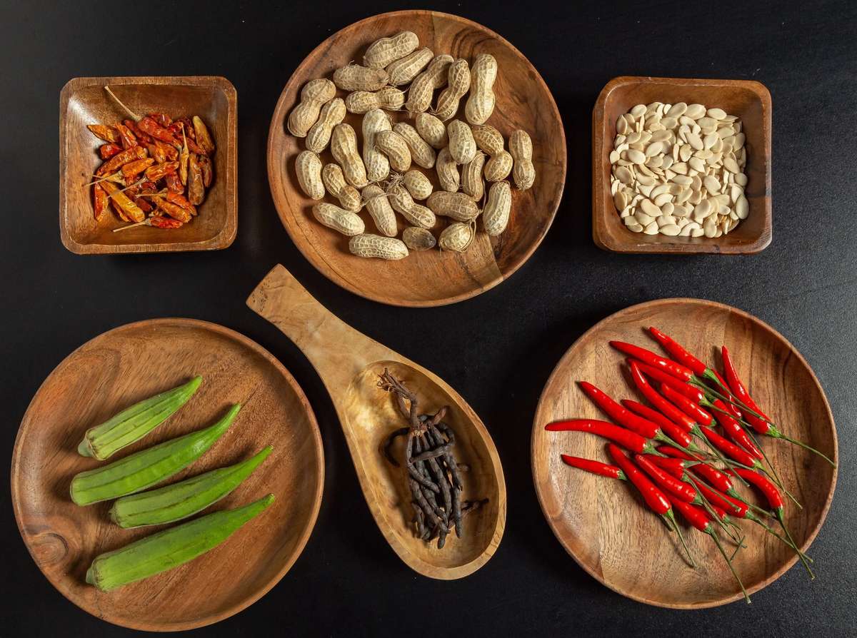 Assorted ingredients in wooden bowls: red chili peppers, peanuts (shelled and unshelled), dried chilies, okra, and dried spice pods.