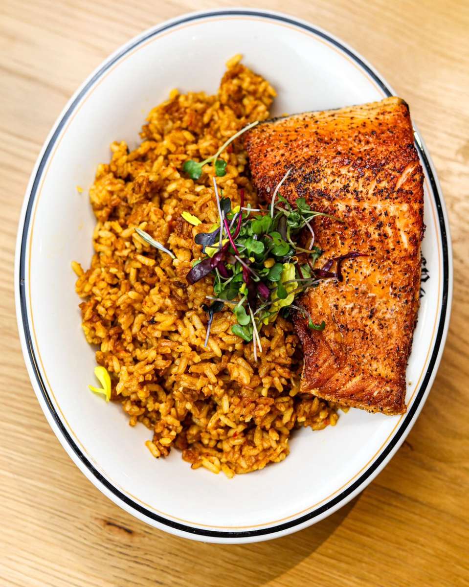 Spiced jollof rice served with a seared salmon fillet and microgreens in a white oval bowl.
