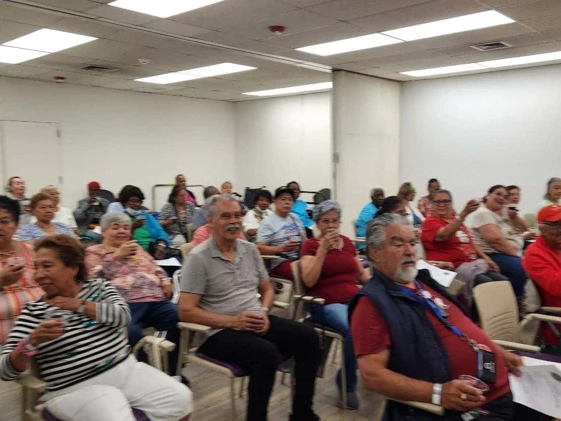 A diverse group of older adults seated in a brightly lit room, attentively participating in a community or educational event.