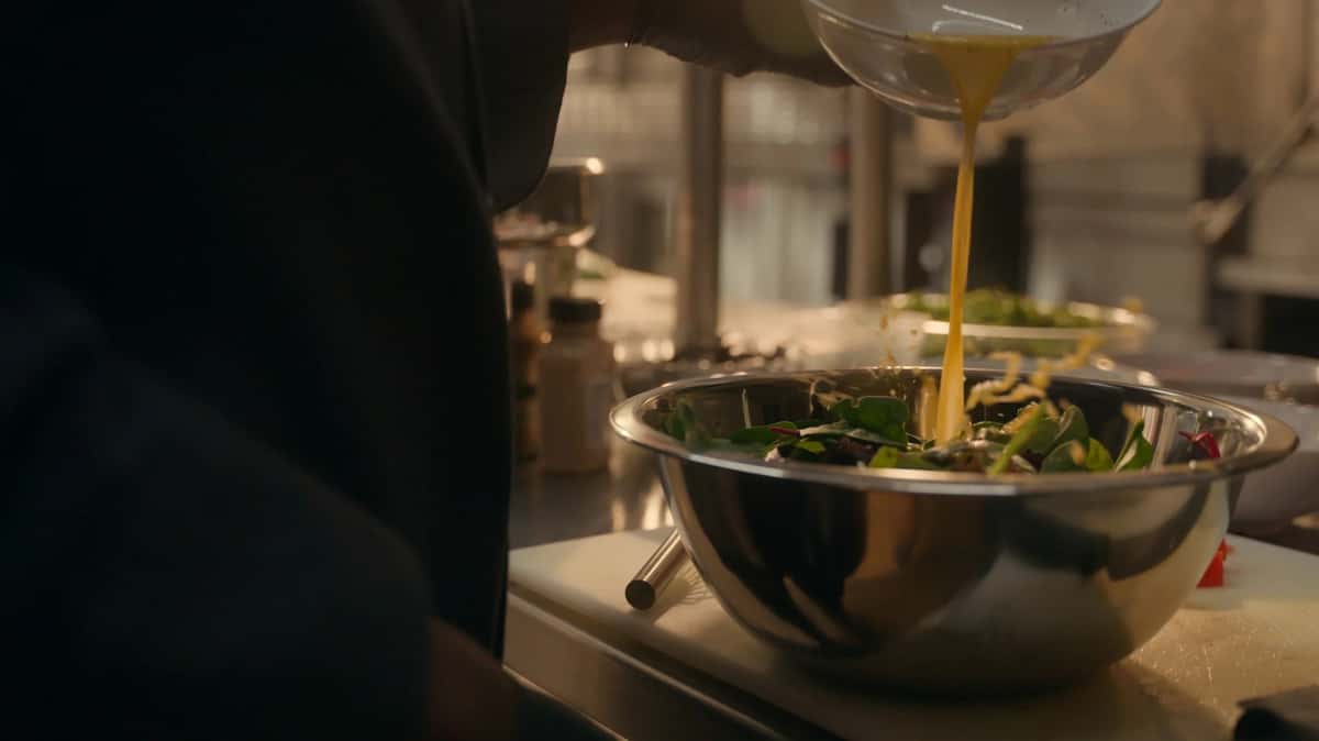 Chef pouring dressing over a fresh green salad in a stainless steel mixing bowl on a kitchen counter.