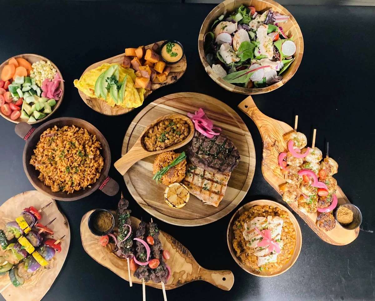 Colorful spread of grilled meats, rice, salads, and vegetable skewers arranged on wooden boards and bowls against a black background.