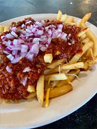 Chili cheese fries topped with diced red onions, featuring golden French fries smothered in melted cheddar cheese and hearty beef chili, served hot on a white diner-style plate.