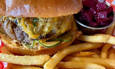 Close-up of a gourmet cheeseburger loaded with melted cheddar, caramelized onions, and fresh zucchini on a toasted brioche bun, served with golden French fries and a side of pickled beets ā classic American comfort food from The Riders Club CafĆ©.