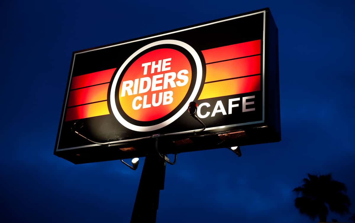 Illuminated roadside sign for The Riders Club Cafe, showcasing a bold retro design with gradient red and orange stripes, and white text on a circular red background, set against a deep blue evening sky.