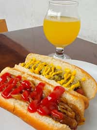 Close-up of two classic hot dogsāone topped with grilled onions and ketchup, the other with relish and mustardāserved with a glass of light, golden craft beer at The Riders Club CafĆ©.