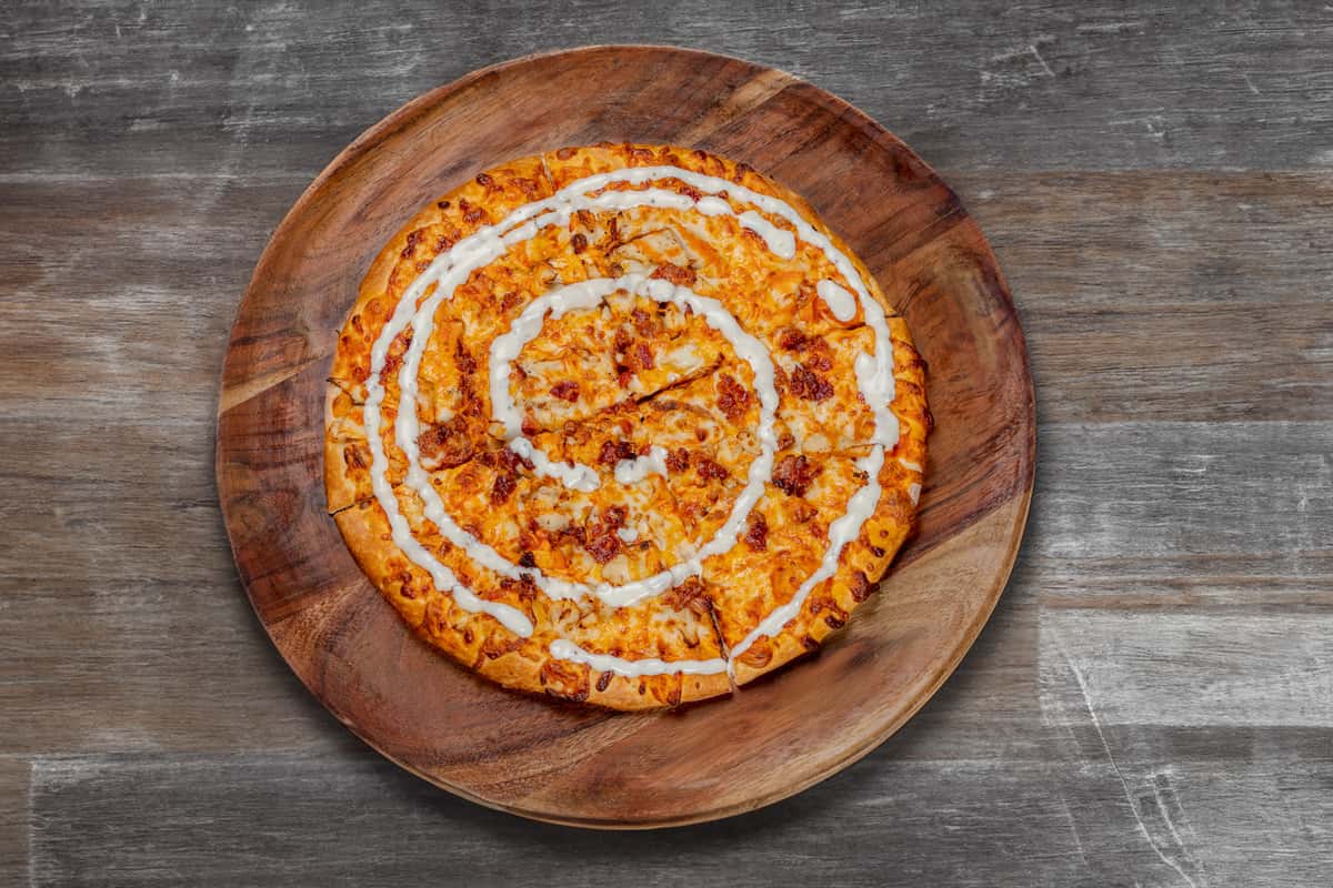 Buffalo chicken pizza topped with melted cheese, diced chicken, and a spiral drizzle of creamy ranch, served on a wooden platter.