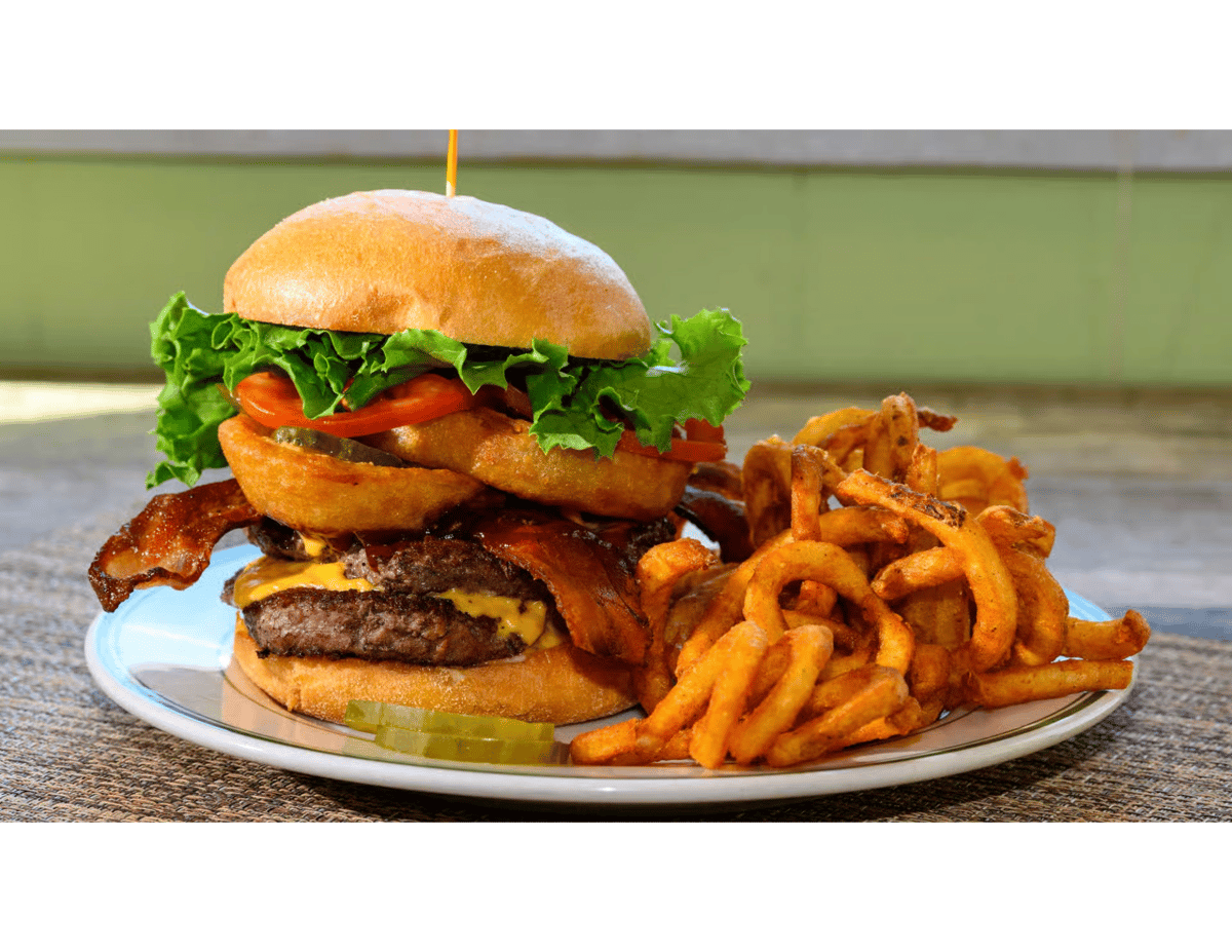 Bacon cheeseburger stacked with onion rings, lettuce, tomato, and pickles, served with crispy curly fries on a white plate.