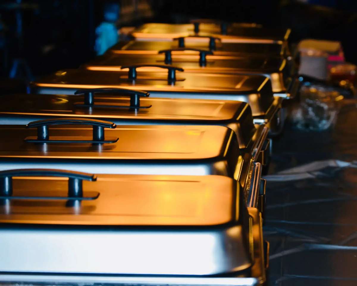 Row of stainless steel catering chafing dishes with lids, lined up on a table, reflecting warm golden light.