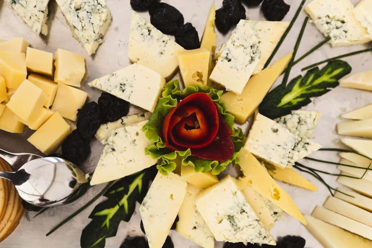 Assorted cheese platter with blue cheese, cheddar cubes, dried prunes, and a decorative vegetable rose garnish in the center.
