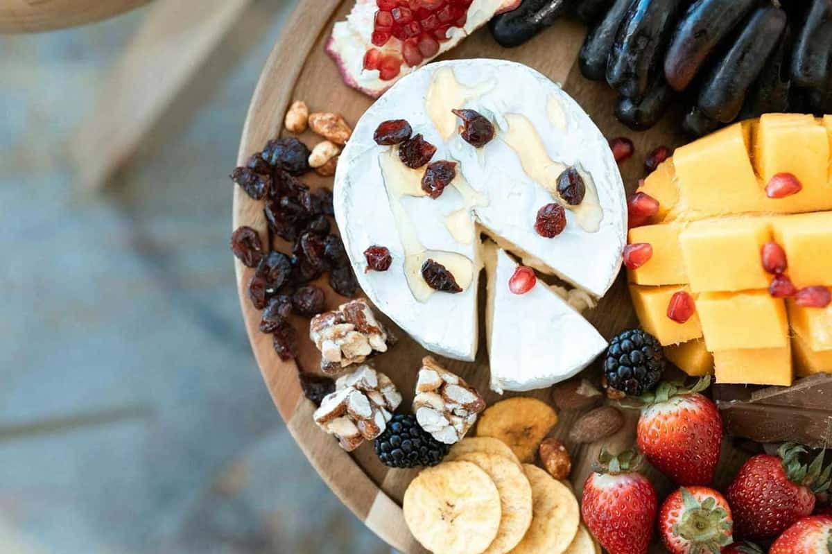 Cheese platter with brie, cheddar, fresh strawberries, blackberries, grapes, dried fruits, nuts, and banana chips garnished with pomegranate.