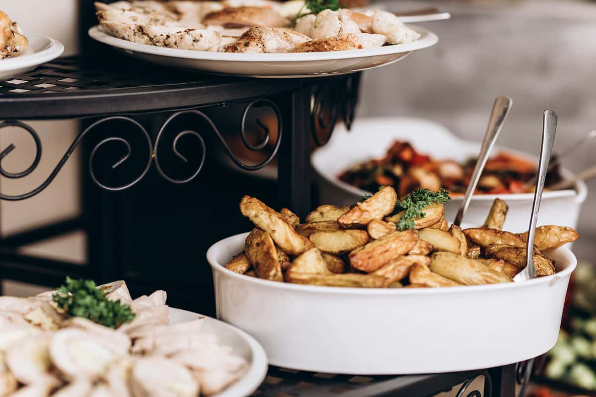 Buffet spread with golden roasted potato wedges, grilled chicken, and vegetable dishes served in white bowls and platters.