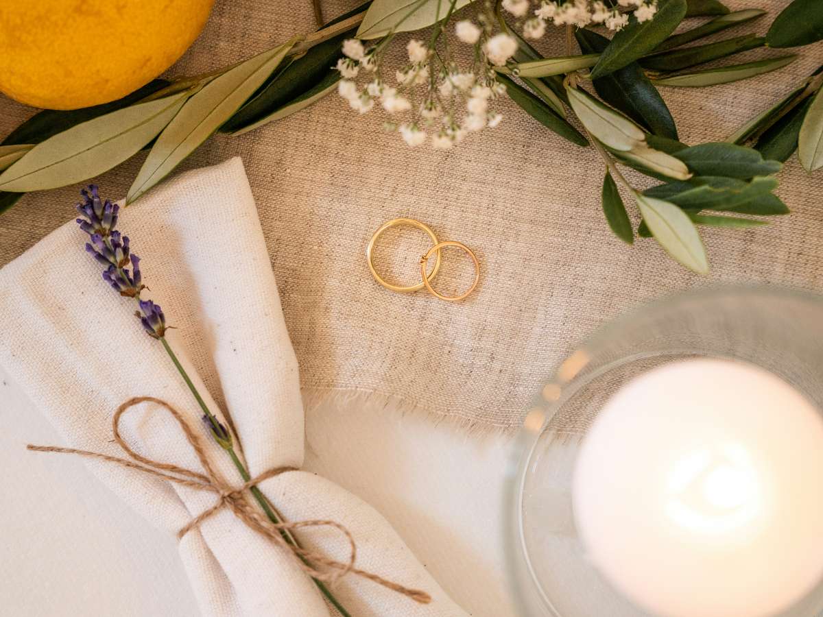 Two gold wedding rings placed on linen with greenery, a tied napkin with lavender, and a candle creating a romantic setting.