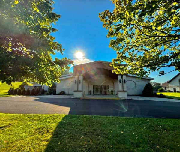 Banquet hall exterior with a covered entrance, framed by trees and bright sunlight casting across the driveway and lawn.