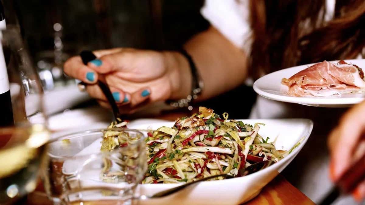 Person serving food at a dining table with a fresh mixed salad in a white bowl and a plate of cured meat slices.