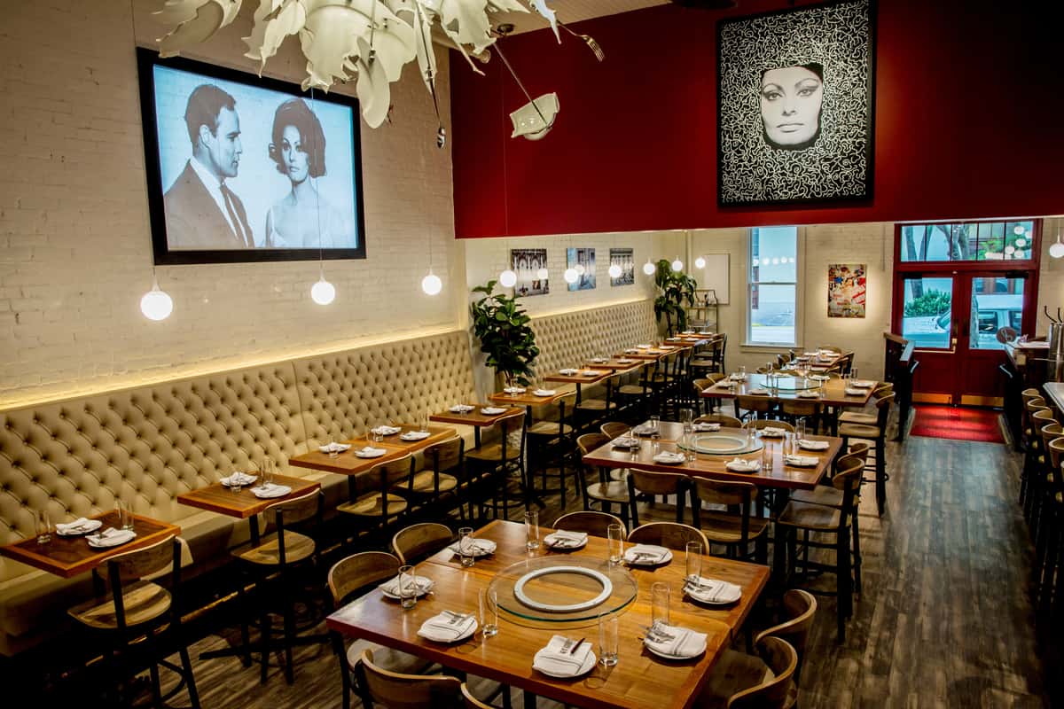 Interior of Sofia restaurant in New Orleans, featuring elegant tufted banquette seating, wooden tables set with glassware and white napkins, modern pendant lighting, and bold artwork including a black-and-white portrait of Sophia Loren on a red accent wall.