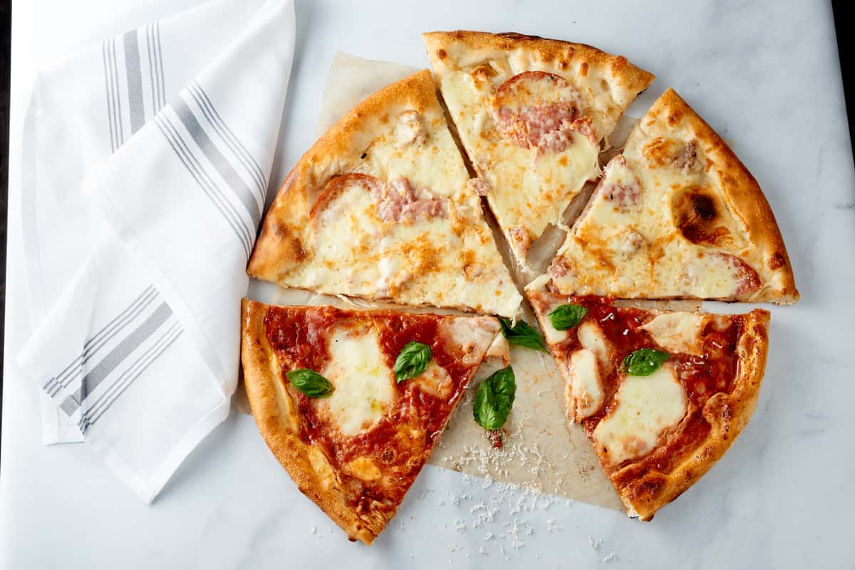 Overhead view of a wood-fired pizza with both red and white sauce slices, topped with mozzarella, fresh basil, and cured meats, served on parchment paper with a striped white napkin on a marble surface.