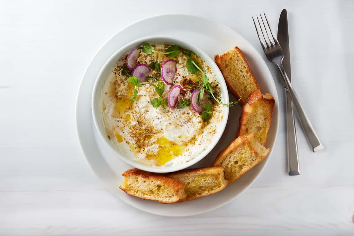 A bowl of housemade whipped ricotta drizzled with olive oil and garnished with thinly sliced radishes, herbs, and spices, served with toasted slices of golden focaccia on a white plate with fork and knife.