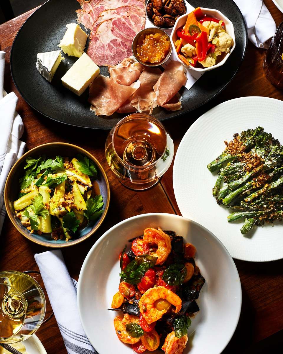 This vibrant overhead shot showcases a delicious spread from Sofia NOLA, featuring a charcuterie board with artisan cheeses, cured meats, and pickled vegetables, alongside seasonal small plates like grilled broccolini, zucchini salad, and squid ink pasta with shrimp. Glasses of white wine complete the inviting, family-style Italian dining experience in the heart of New Orleans.